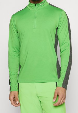 Homme portant un haut de sport à manches longues vert vif avec des panneaux latéraux gris foncé et un pantalon vert assorti, debout devant un fond blanc.