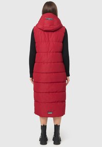 Manteau sans manches rembourré rouge avec une capuche, présentant des sections matelassées, un design long et un logo au dos. Porté par-dessus un haut noir à manches longues.
