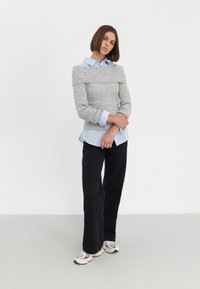 Maglione grigio off-the-shoulder con texture a costine indossato sopra una camicia button-up azzurro chiaro, abbinato a pantaloni neri a gamba larga e sneakers multicolori.