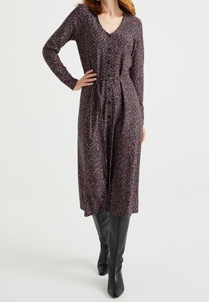 Kobieta ubrana w czarną, długorękawową sukienkę midi z drobnym kwiatowym wzorem, z guzikami z przodu oraz czarne, skórzane kozaki za kolano.