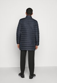 Marinblå pufferjacka med horisontell quiltning, hög krage och avslappnad passform. Buren över svarta byxor och skor med vit sula.