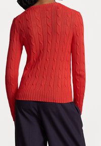 Frau trägt einen leuchtend roten Pullover mit Kabelstrickmuster und lockere dunkle Hose, von hinten dargestellt vor einem schlichten weißen Hintergrund.