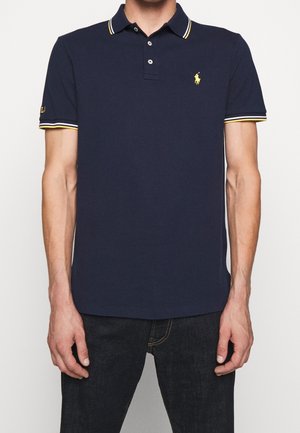 Granatowa koszulka polo z klasycznym kołnierzykiem, żółte i białe paski na rękawach oraz małe haftowane logo na piersi.