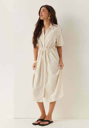 Femme aux longs cheveux foncés portant une robe crème boutonnée longueur genou avec une taille à cordon, des sandales noires, des boucles d'oreilles étoile de mer et un bracelet blanc.