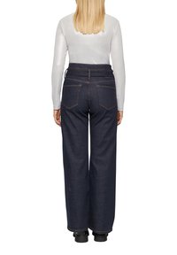 Dunkelblaue weit geschnittene Jeans mit hoher Taille, hellem Nähten und zwei Gesäßtaschen, aus Denimstoff. Kombiniert mit einem langärmeligen, metallischen Oberteil.