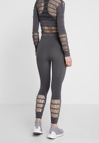 Šedý sportovní top s vystřiženým síťovým vzorem, doplněný o přiléhavé šedé legíny s horizontálními otvory na dolní části nohy a elastickým pásem se značkou.