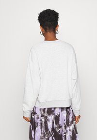 Personne aux cheveux courts et bouclés portant des boucles d'oreilles anneaux en argent, un sweat-shirt gris clair et une jupe à motifs violets et noirs, vue de dos.