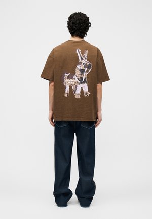 Uomo con capelli ricci che indossa una t-shirt marrone oversize con una grande immagine di una lama stilizzata sul retro, abbinata a jeans scuri larghi.