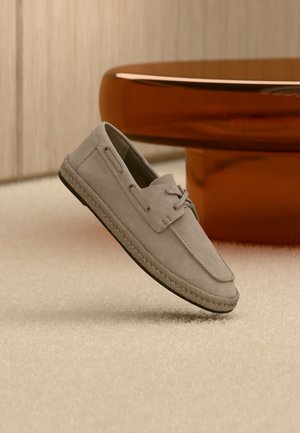 Lichtbruine suède bootschoen met veterdetailing en espadrille-achtige zool, rechtop geplaatst op een beige tapijt naast bruin rond meubelstuk.