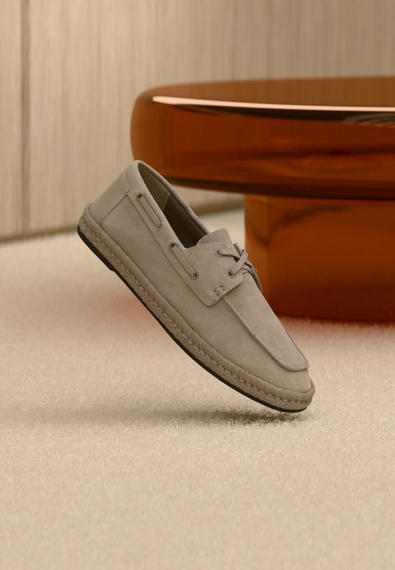 Chaussure bateau en daim marron clair avec détails en lacets et semelle style espadrille, positionnée à la verticale sur un tapis beige près d'un meuble circulaire marron.