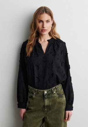 Femme aux cheveux châtain clair portant un chemisier noir texturé et un pantalon taille haute vert olive, debout devant un mur gris clair uni.