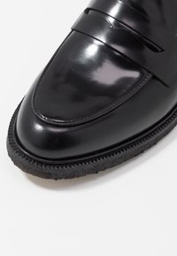 Gros plan d'une chaussure mocassin en cuir noir poli, avec un bout arrondi et une semelle épaisse texturée, sur fond blanc.