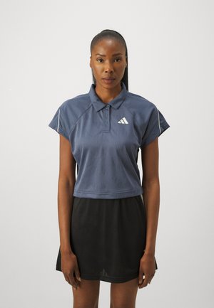 Marineblaues, kurzärmliges Poloshirt mit verkürztem Schnitt, strukturiertem Stoff und weißem Logo auf der Brust, kombiniert mit einem schwarzen Rock.
