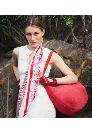 Frau in weißem Kleid und floralem Schal hält große rote Handtasche und passende fingerlose Handschuhe, steht vor Felsen und Pflanzen.