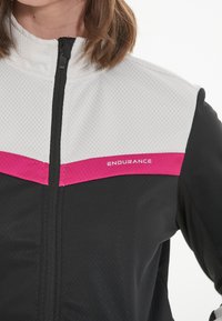 Schwarze Sportjacke mit einem weißen oberen Abschnitt im Karomuster, pinken Akzenten und einem Frontreißverschluss. Branding auf der Brust.