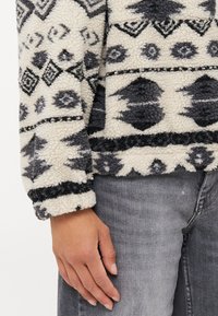 Pull en polaire avec une base crème et des motifs géométriques gris foncé, surface texturée et poignets côtelés. Associé à un jean gris.