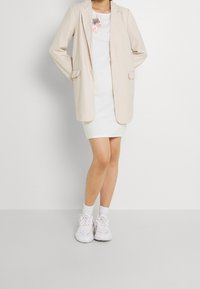 Beżowa marynarka o luźnym kroju, nałożona na białą sukienkę z kwiecistym haftem, dopełniona białymi trampkami i skarpetkami do kostek.