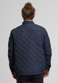 Marineblauwe gewatteerde jas met een opstaande kraag, met diamandpatronen en drukknopen, vervaardigd uit een gestructureerde, lichte stof.