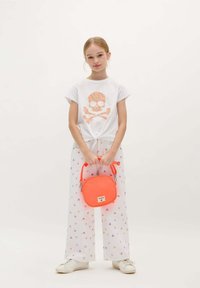 White t-shirt with a colorful skull and crossbones design, paired with patterned sheer wide-leg pants and a small neon orange handbag.