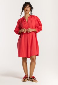 Koraalfarbenes kurzes Kleid mit rundem Ausschnitt und Ballonärmeln. Kombiniert mit schwarzen Sandalen mit floralen Akzenten. Glatte, leichte Stoffqualität.