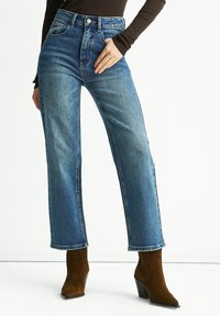 High-waisted blue denim jeans with a straight cut, featuring front pockets and a button closure. Paired with brown heeled ankle boots.