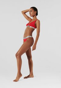Crveni bikini set od glatkog materijala, s trokutastim vrhom i donjim dijelom s niskim strukom koji ima srebrne logotip trake duž rubova.