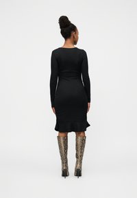 Μαύρο μακρυμάνικο φόρεμα με φαρδιά βάση, σε συνδυασμό με ψηλές μπότες στιλέτο με σχέδιο φίδι, από πίσω όψη. Λεία υφή, εφαρμοστό σχέδιο.