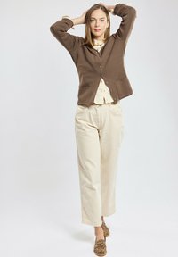Cardigan marrone sopra una camicia leggera con bottoni, abbinato a pantaloni crema e scarpe slip-on con stampa leopardata. Il cardigan ha bottoni e tasche.