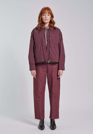 Vrouw met rood haar die een bordeauxrode jas, bijpassende broek en zwarte laarzen draagt, staand tegen een effen lichtgrijze achtergrond.