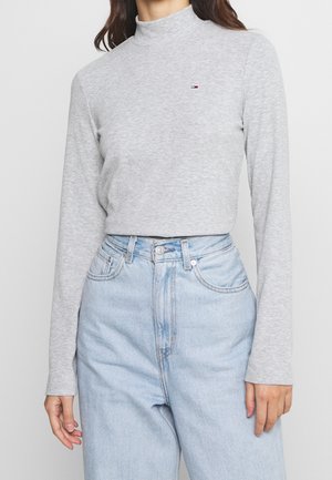 Femme portant un pull à col roulé côtelé gris clair à manches longues et un jean en denim bleu clair taille haute, debout devant un fond uni.