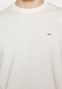 Krémové bavlněné tričko s kulatým výstřihem a malým černým logem "W.W." na levé straně hrudi. Měkká textura, uvolněný střih.