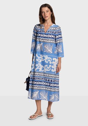 Kafethán azul con patrones en varios tonos de azul, diseños geométricos y florales, mangas hasta tres cuartos, largo hasta la rodilla, llevado con sandalias.