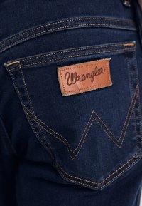 Tumed sinised teksad sisaldavad pruunist nahast sildi, millel on hõbedaste õmblustega vöökoht ja kus on kiri "Wrangler" kalligraafiliselt. Silmapaistev oranž õmblus ja iseloomulik tasku kujundus.