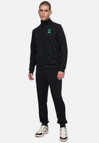 Veste de survêtement zippée noire avec un col haut et un logo vert, accompagnée d'un pantalon assorti. Le tissu semble lisse et confortable, adapté pour les vêtements de sport.