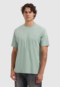 Jonge man met krullend haar, in een lichtgroen T-shirt met de tekst "PURE PATH", staand tegen een effen lichtgrijze achtergrond.