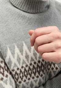 Hand in een losse vuist dichtbij de borst, draagt een grijs gebreid trui met een ronde boord.