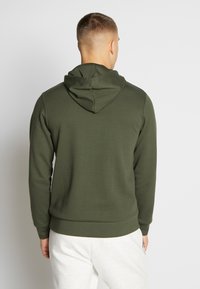 Felpa verde con finitura testurizzata, cappuccio con coulisse regolabile e polsini e orlo a coste. Indossata con pantaloni della tuta grigi chiari.