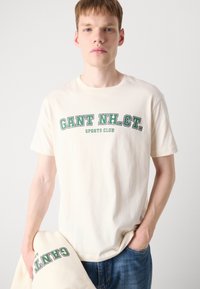 Cream-colored cotton T-shirt featuring "GANT NH. CT. SPORTS CLUB" text in green. Short sleeves, crew neckline, relaxed fit, held by a model.