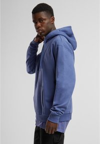 Felpa blu con cappuccio e tasca frontale, caratterizzata da una vestibilità rilassata e polsini a costine. Texture liscia con un aspetto a strati all'orlo.