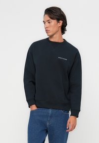 Kék crewneck pulóver pamut keverékből, bordás mandzsettával és aljjal, a mellkason egy kis fehér logóval. Lazán illeszkedő, minimális dizájn.