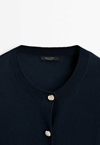 Cardigan en maille bleu marine avec un col rond, présentant deux boutons décoratifs argentés et une étiquette de marque visible au niveau du col.