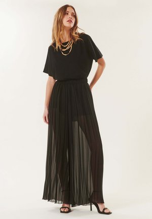 Femme portant un haut noir à manches courtes, des colliers dorés superposés, un pantalon noir large plissé transparent et des sandales noires à talons hauts, debout.