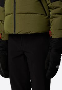 Zöld puffernadrág rácsos mintával és fekete kiegészítőkkel, fekete nadrággal és logós fekete kesztyűkkel párosítva. Piros cipzárhúzók láthatók.