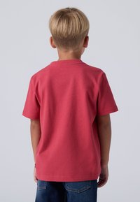 Raudona medvilninė marškinė su trumpomis rankovėmis, apvaliu iškirpimu ir lygiu tekstūru, rodomas iš nugaros. Derinamas su mėlynomis džinsinėmis kelnėmis.