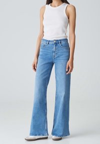 Femme portant un jean large bleu clair, un haut sans manches blanc et des chaussures blanches à enfiler avec semelles marron, debout devant un fond uni.