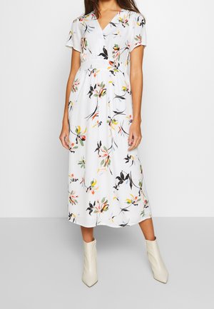 Vrouw draagt een witte midi-jurk met kleurrijke bloemenprints, korte mouwen, V-hals en witte enkellaarzen, staand tegen een effen achtergrond.