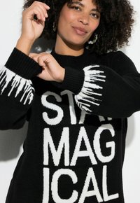 Suéter negro de punto con letras blancas audaces "STAY MAGICAL" en el frente, con detalles texturizados de flecos en las mangas.