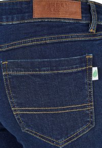 Donkerblauwe denim jeans met contrasterende gele stiksels en een klein groen label. Bevat een standaard achterzakontwerp met leren etiket.