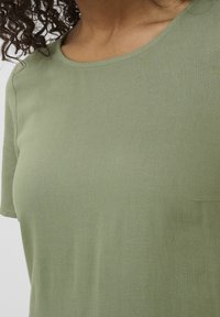 Vrouw in een lichtgroen shirt met korte mouwen en een ronde halslijn, weergegeven van de schouders tot het midden van de torso.