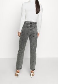 Jeans de mezclilla oscuros de tiro alto con piernas rectas, que cuentan con dos bolsillos traseros y detalles de costura en contraste. Combinados con zapatos de tacón blancos.